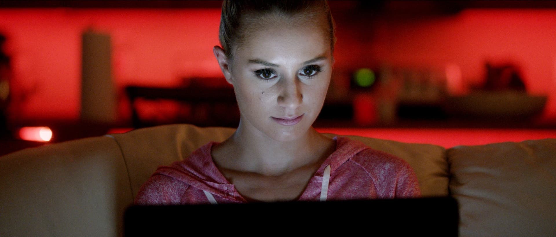 Young woman in pink hoodie focused on laptop in ambient red lighting