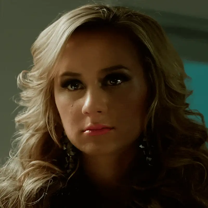 Close-up of a woman with curly hair and dramatic makeup, showcasing her intense expression and earrings.