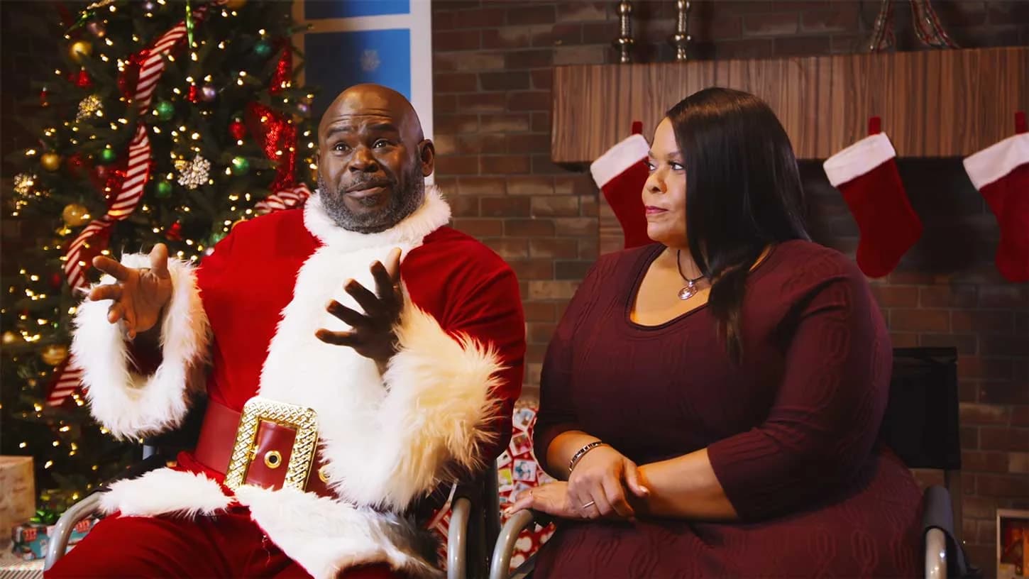 A man in a Santa costume and a woman sitting together in front of a decorated Christmas tree.