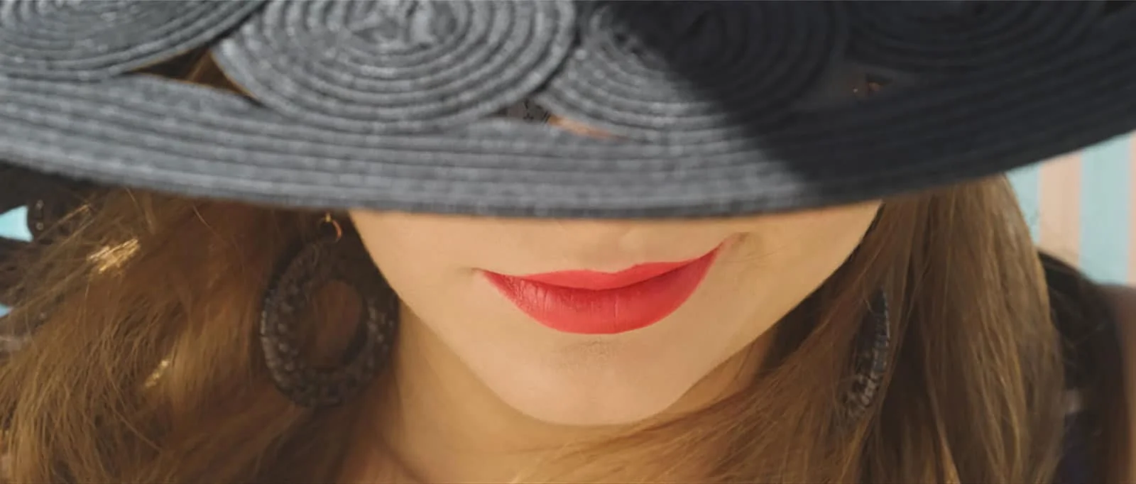 Close-up of a woman's lips with red lipstick, wearing a wide-brimmed black hat and earrings.
