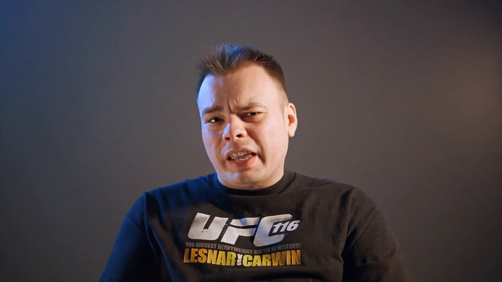 Man wearing a UFC 116 t-shirt, expressing intensity against a dark background.