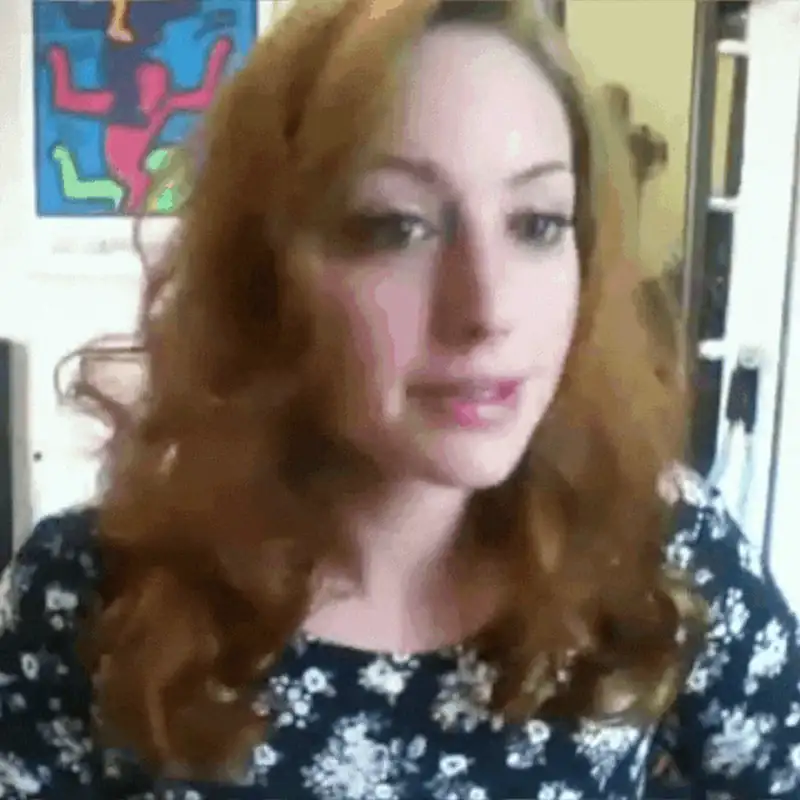 Smiling woman with wavy red hair, wearing a floral dress, in a bright indoor setting.