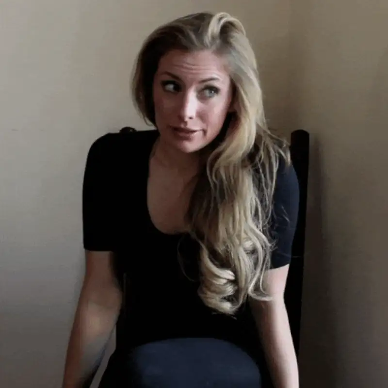Woman with long hair in a black top sitting, looking pensively to the side against a neutral background.