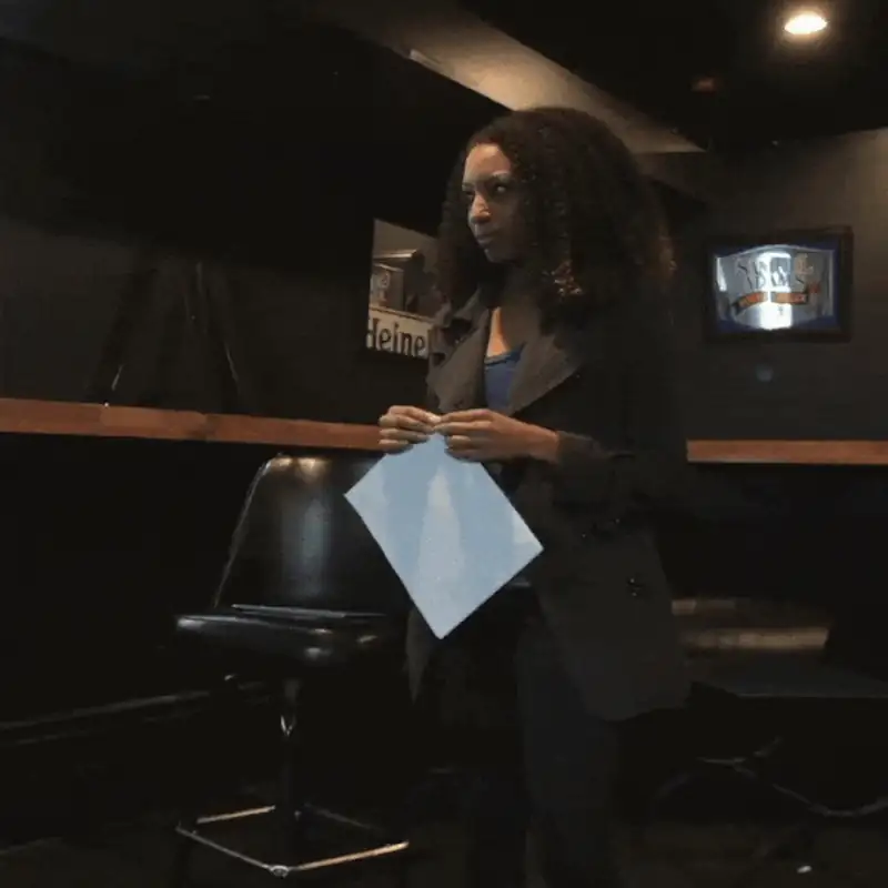 A woman with curly hair stands in a dimly lit bar holding a blank sheet of paper.