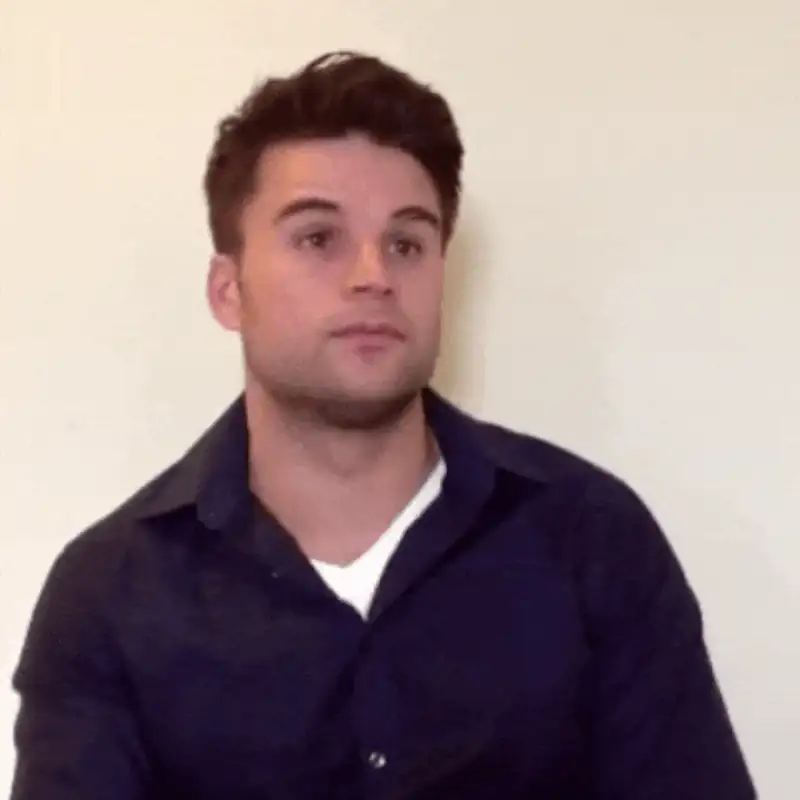 Young man in a navy shirt sitting against a plain background, looking thoughtfully off-camera.