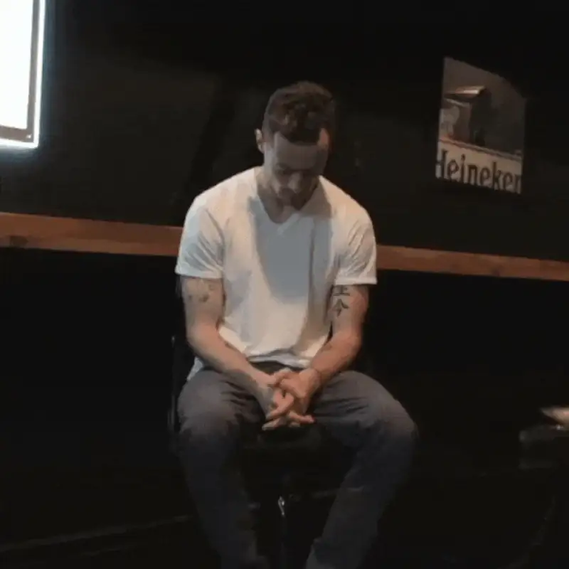 Man sitting on a chair, deep in thought, wearing a white t-shirt and jeans in a dimly lit bar.