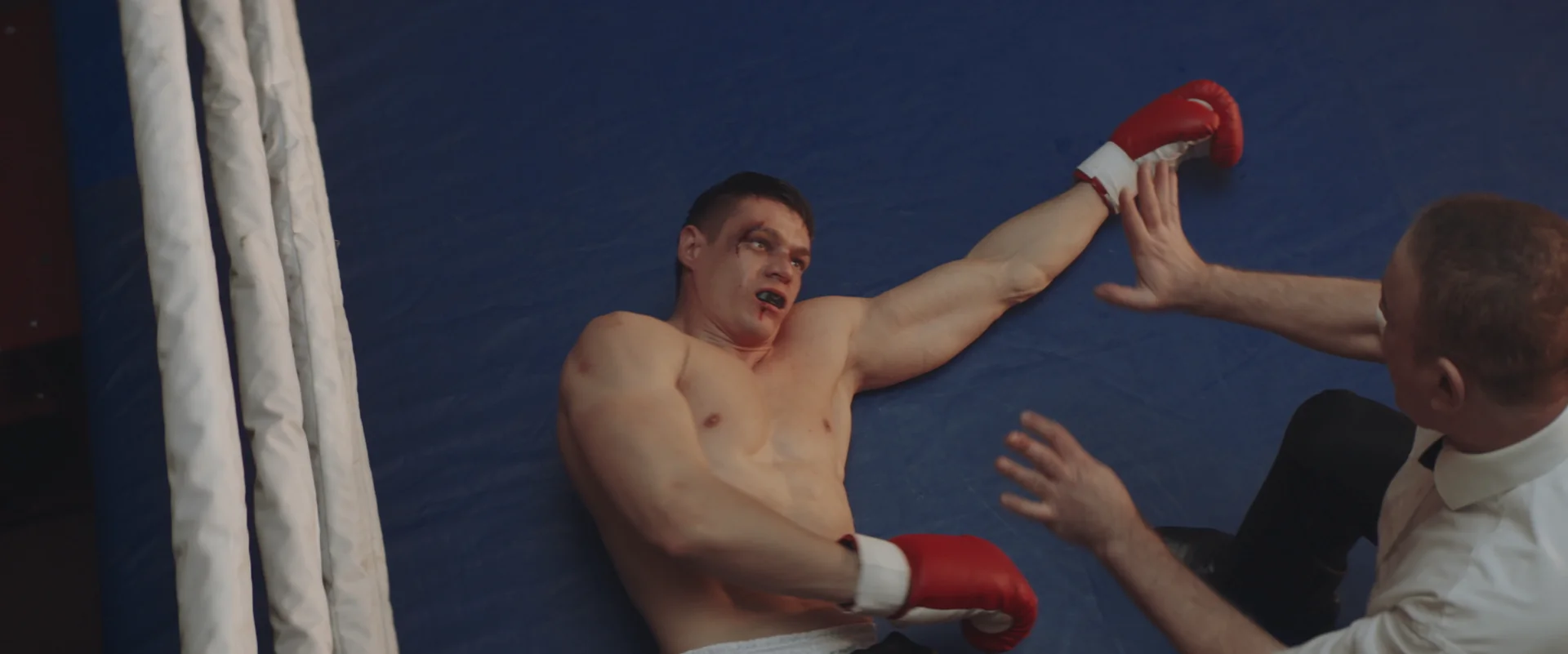 A boxer on the mat with a serious expression and a trainer assisting him.