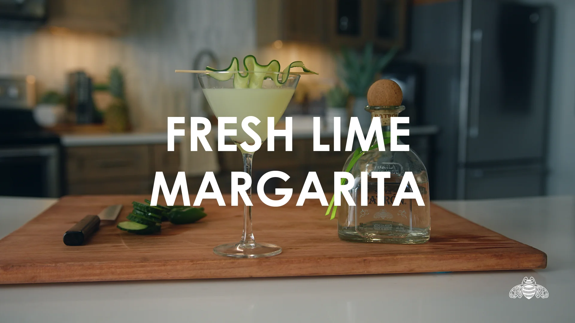 Fresh lime margarita next to sliced cucumbers and a bottle of tequila on a wooden cutting board.