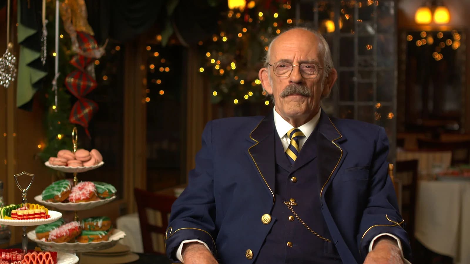An elderly man in a blue suit sits in a festive restaurant setting with dessert platters.