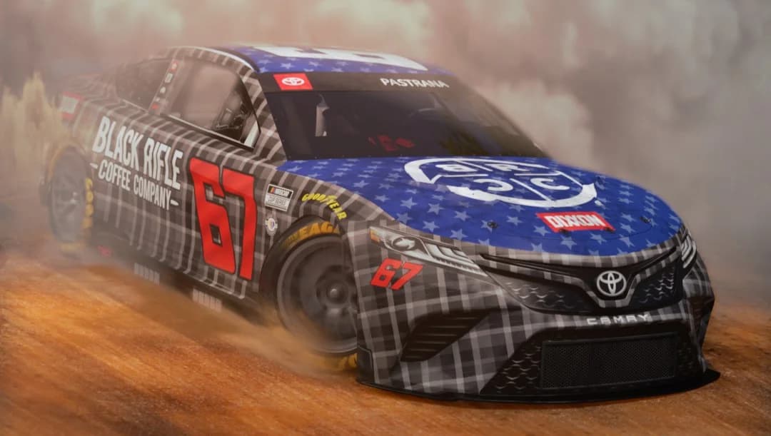 A NASCAR car with Black Rifle Coffee branding skids on a dirt track, kicking up dust.
