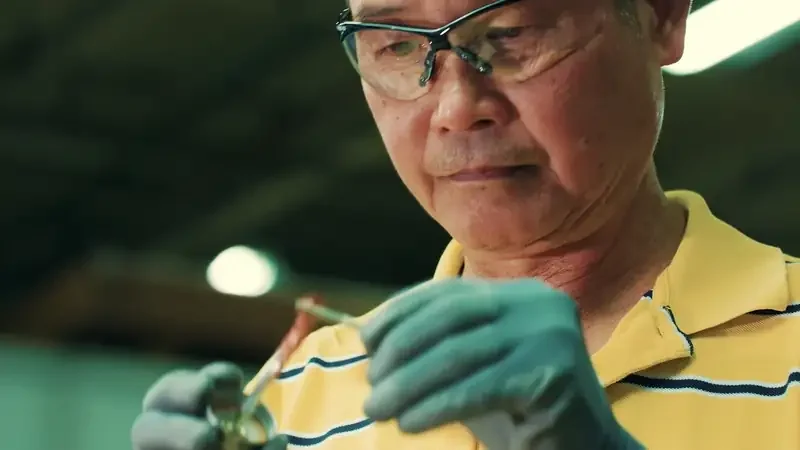 Man in glasses examining a small metal piece in a workshop.