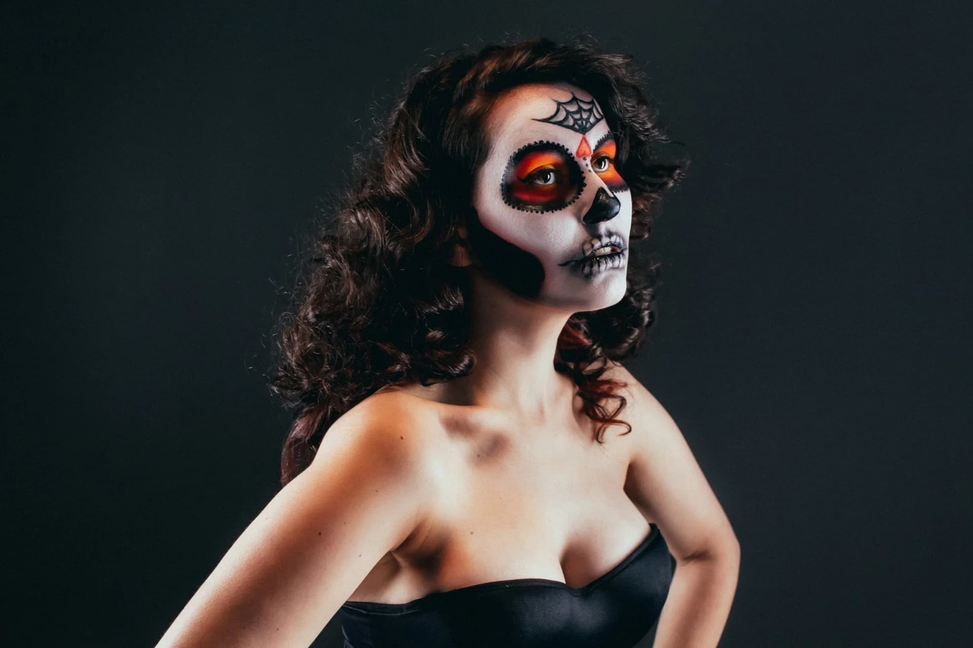 Woman with vibrant sugar skull makeup, wearing a black outfit, posing against a dark background.