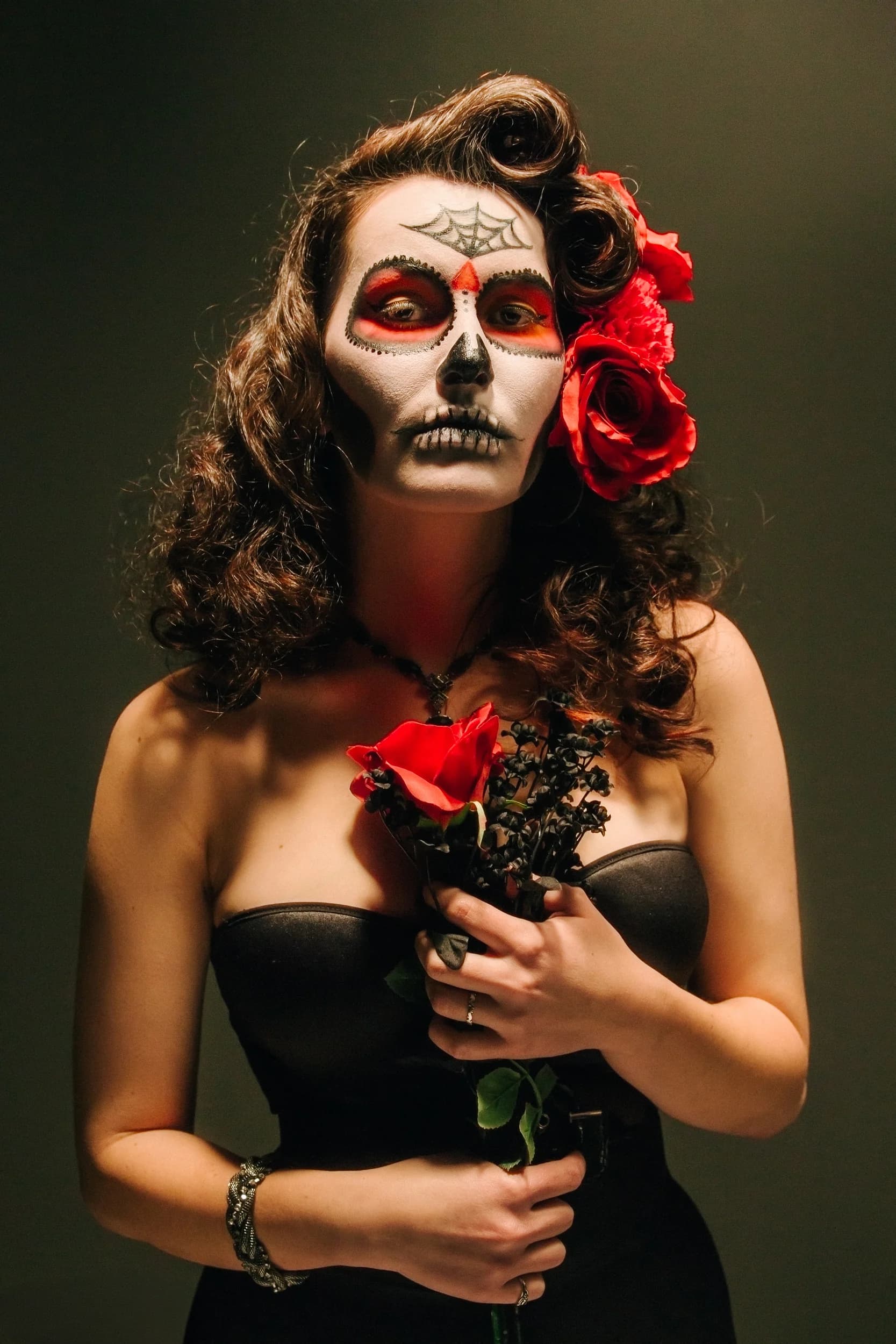 Woman with Day of the Dead makeup holding red roses, wearing a black dress and a dark background.