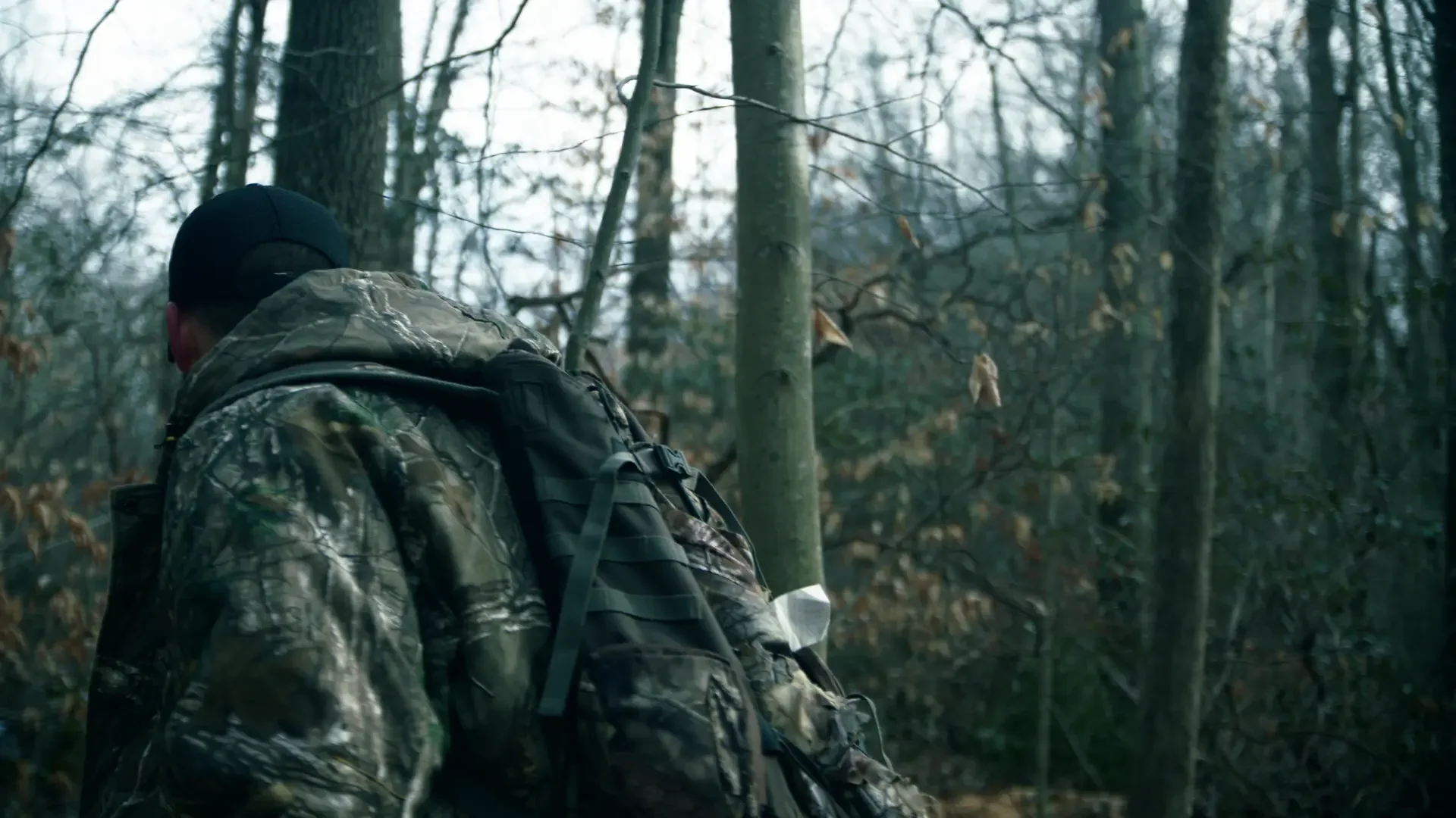 Person in camouflage gear hiking through a forest with bare trees and a rugged landscape.